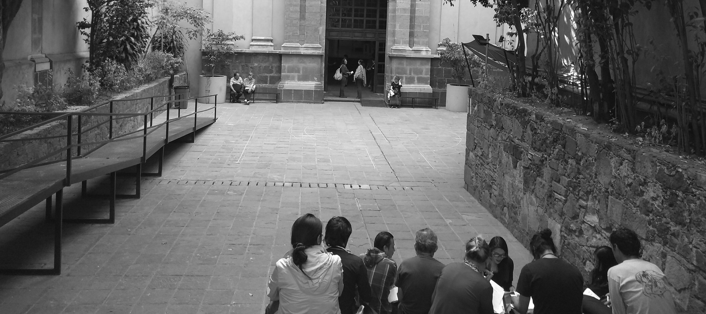 Grupo de personas sentadas en el patio de un recinto cultural, frente a la entrada principal.