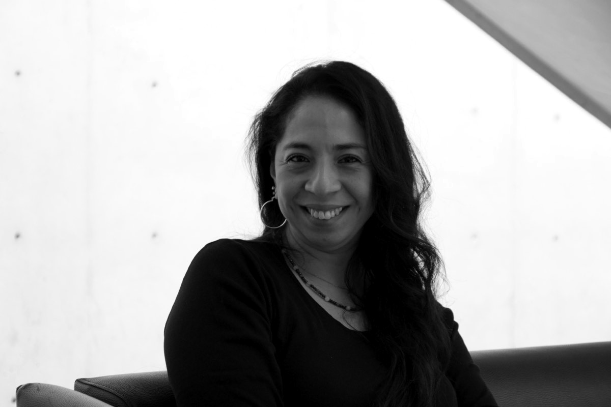 Retrato de Azucena Meza Uscanga sentada, sonriendo, frente a un muro de concreto.